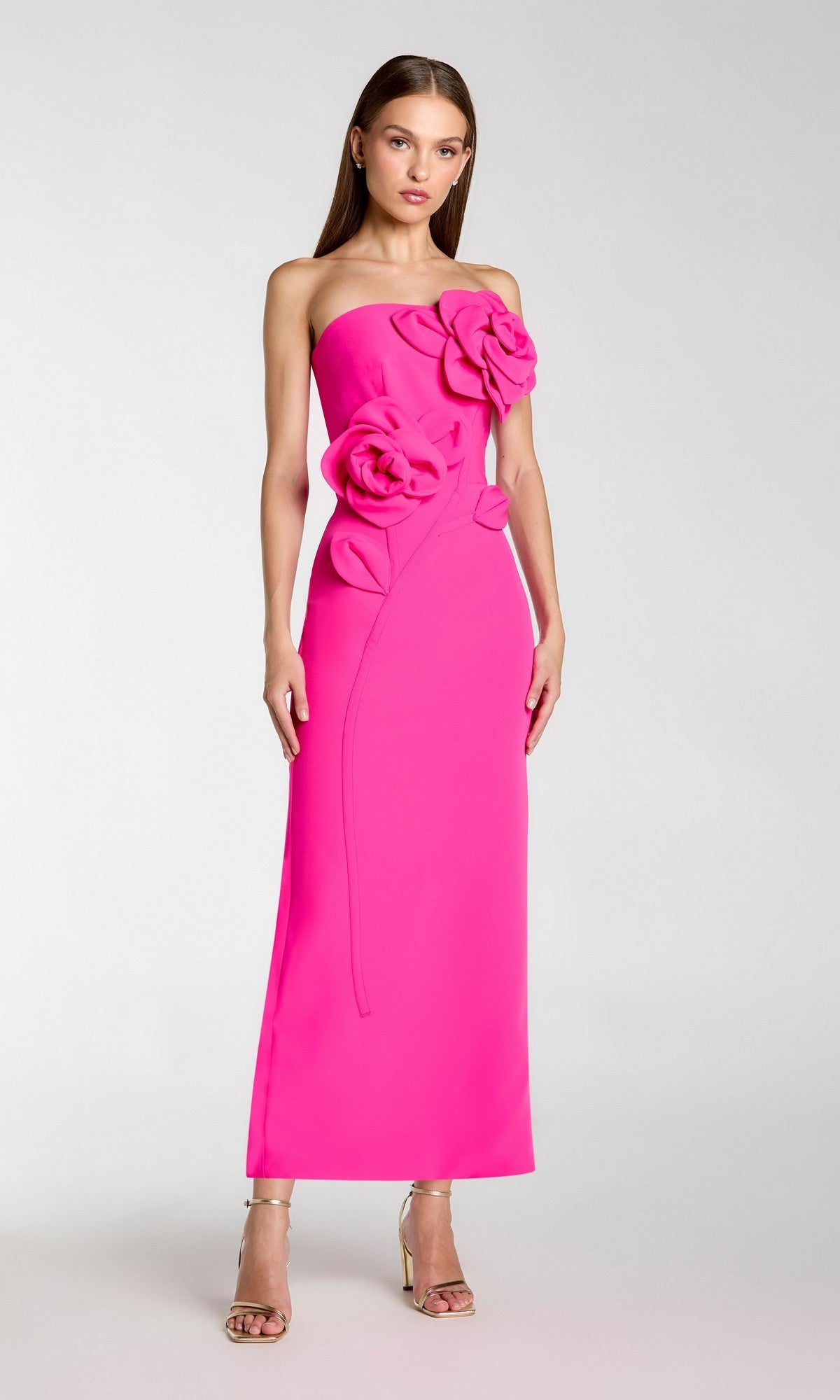 A woman stands against a plain light background wearing the Mac Duggal 2361 Long Formal Dress in hot pink, a strapless crepe midi dress embellished with large 3D floral appliques on the bodice, paired with gold high-heeled sandals.